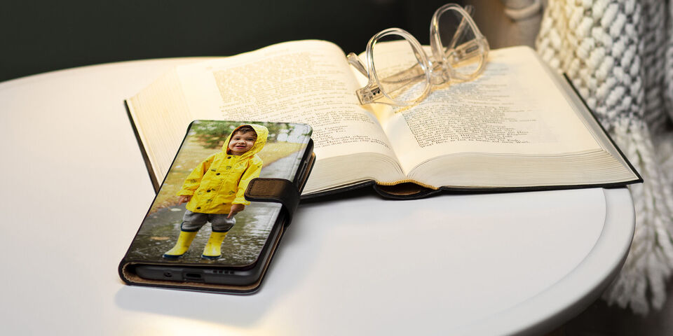 Uno smartphone su un tavolo mostra un bambino felice con un impermeabile. Accanto ci sono un libro aperto e degli occhiali.