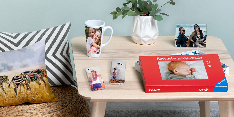 Un tavolo con vari prodotti fotografici: un album CEWE, una tazza con una foto, custodie e un puzzle. Un cuscino a strisce zebrate e piante completano la scena.