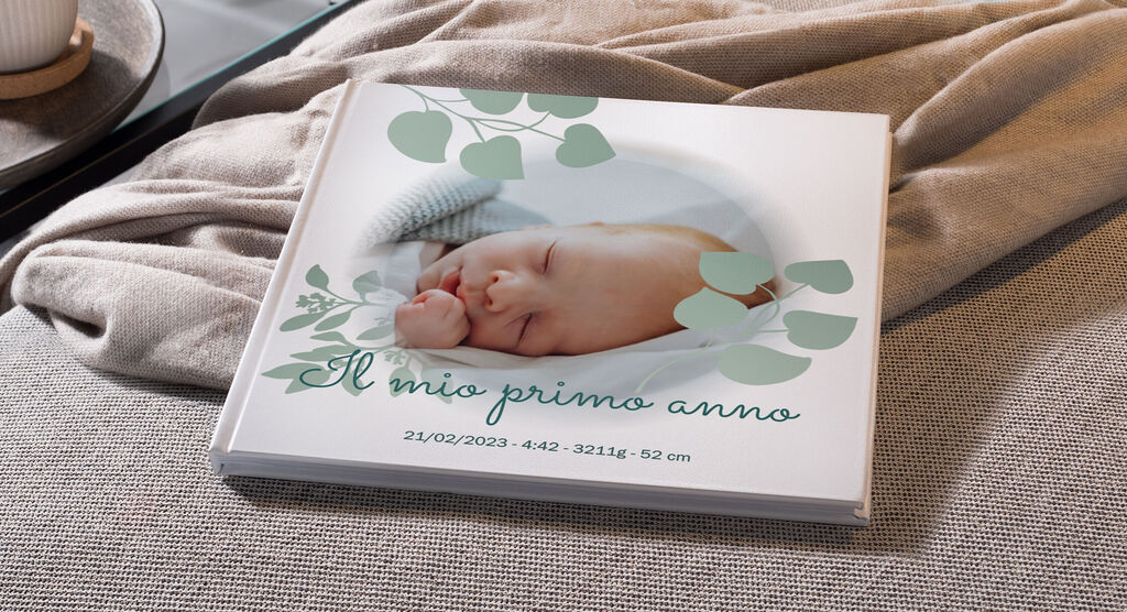 Un bel CEWE Fotobuch con un bambino che dorme su un divano, circondato da foglie tenere. Mostra il titolo "Il mio primo anno".