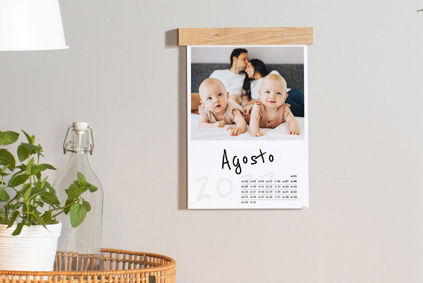 Un calendario da parete è appeso. Il mese di agosto è in evidenza. Due bambini sorridenti sono seduti su una coperta.